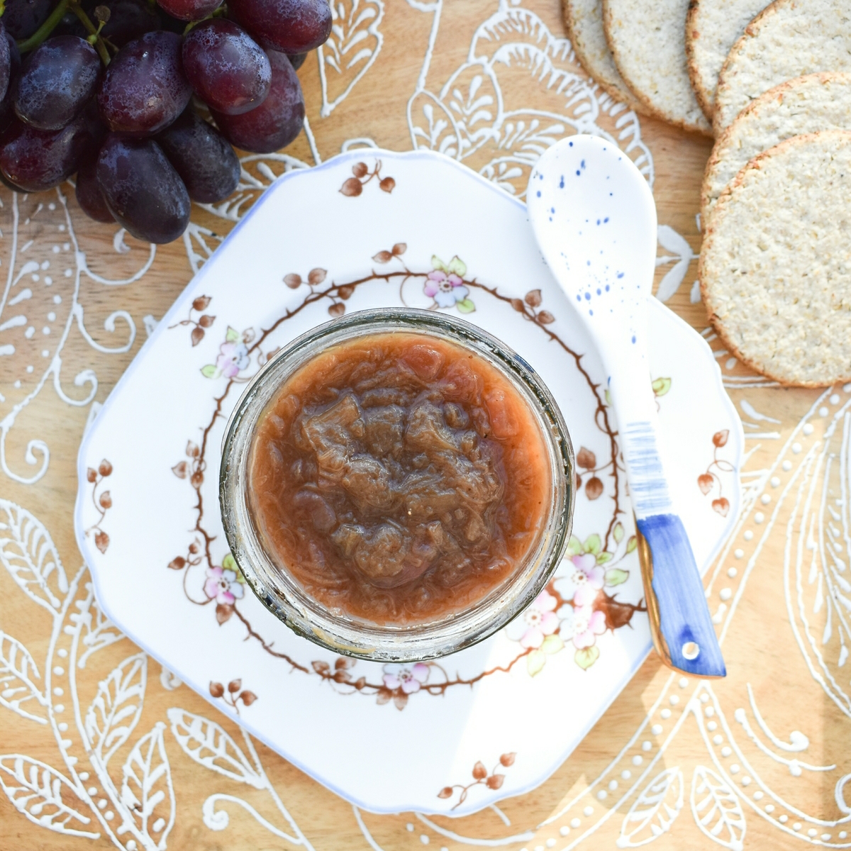 A jar of rhubarb chutney on a wooden tray with grapes and oatcakes.