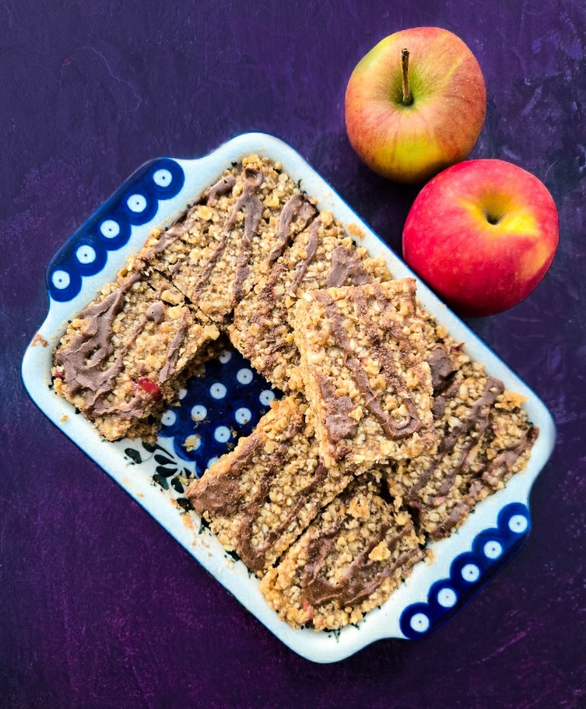 Bars of apple and cinnamon flapjacks in a ceramic dish next to two red eating apples.